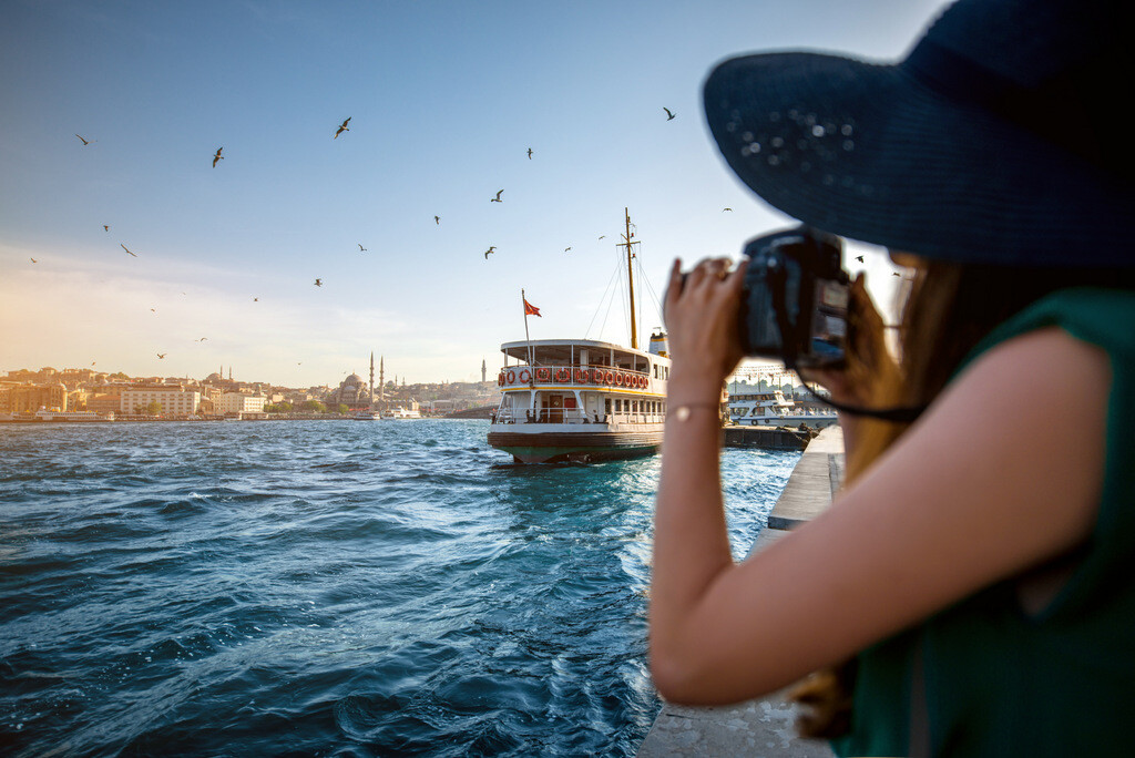 Şapkalı ve fotoğraf makineli turist kadın İstanbul'da Boğaz turu yaparken nostaljik vapurların resmini çekiyor